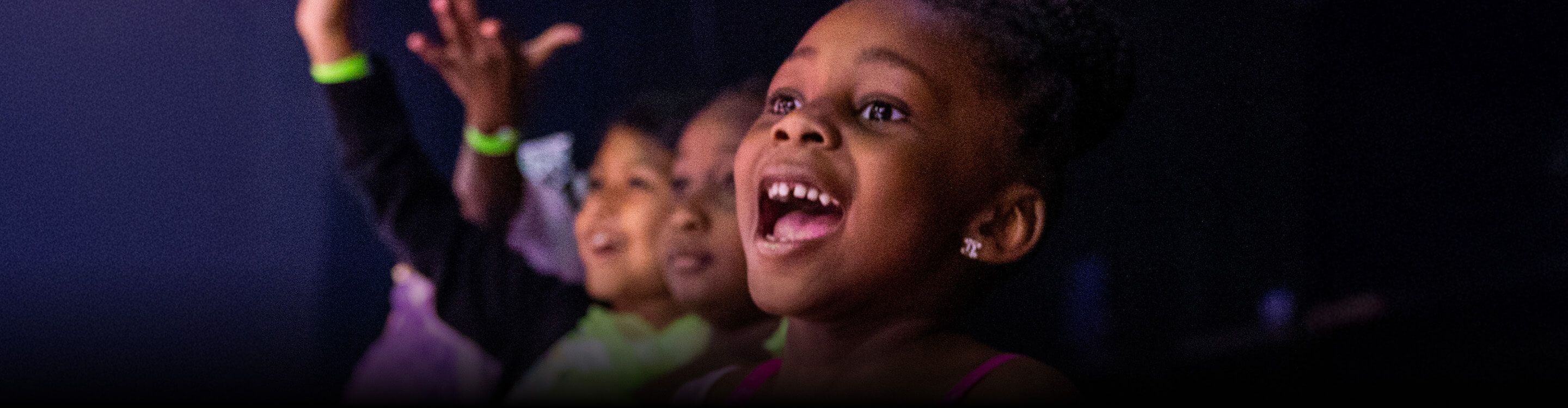 Excited children smiling, cheering, and raising their hands while watching Disney On Ice and Monster Jam performances.